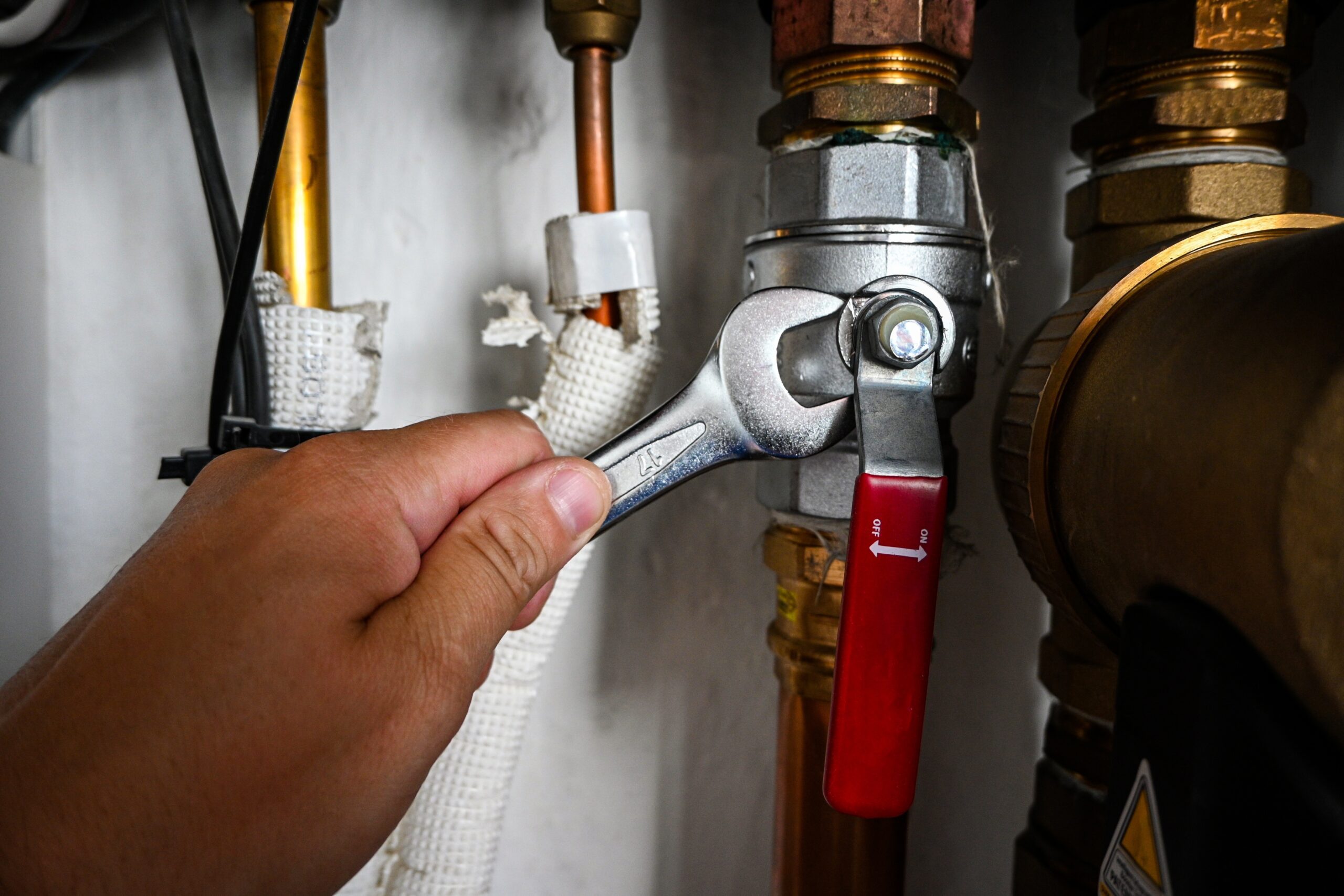 A hand uses a wrench to tighten a metal valve connected to copper pipes and plumbing fittings.